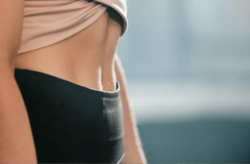 Close-up of a woman's toned midsection wearing a beige crop top and black athletic leggings, showcasing weight loss results.