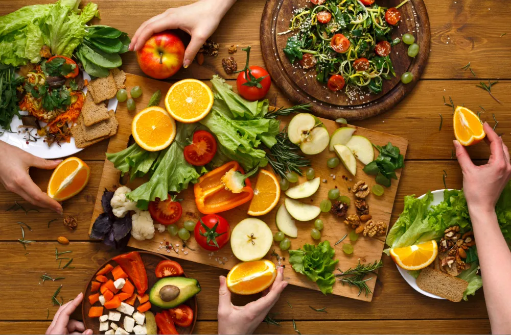 Overhead view of a wooden table with a colorful spread of fresh fruits and vegetables including oranges, apples, tomatoes, and leafy greens, with multiple hands reaching in to grab food.