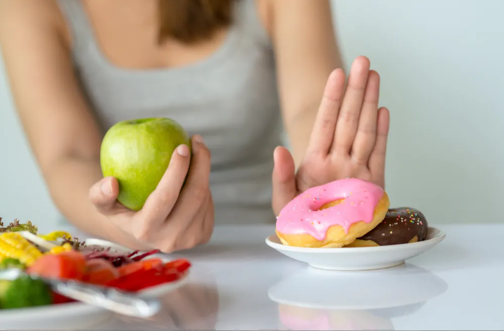 Woman holding a green apple in one hand and pushing away a plate of donuts with the other, choosing healthy food over sweets.