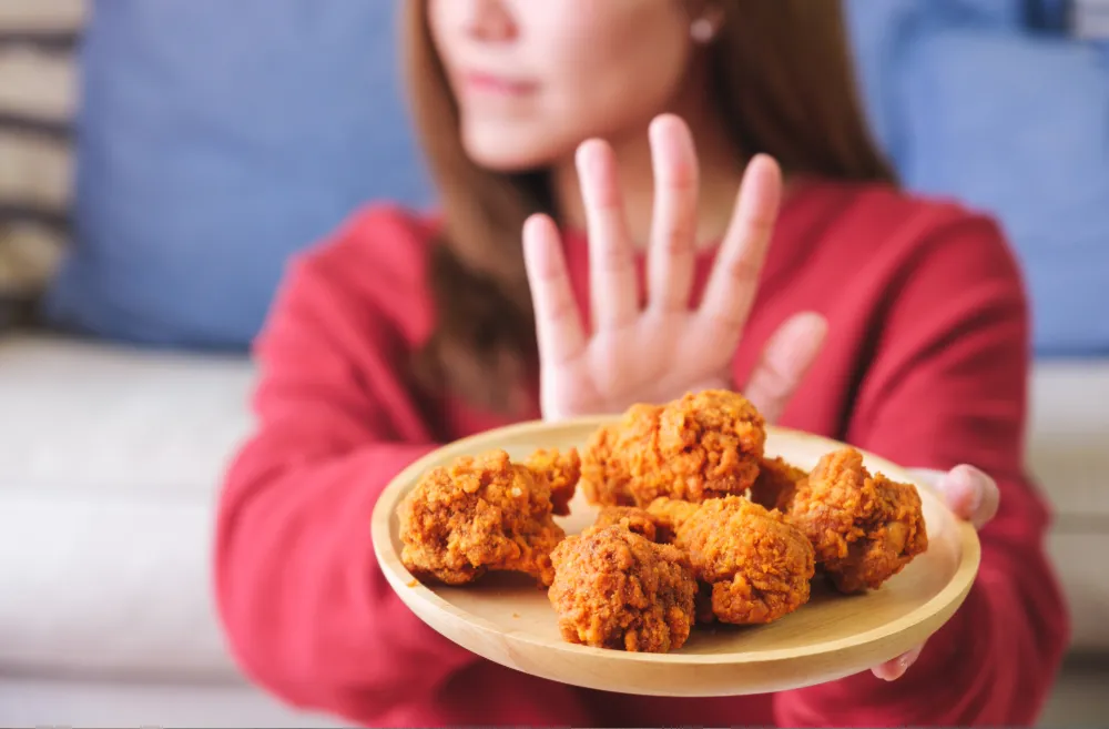 Woman in a red sweater holding a plate of fried chicken while raising her hand to decline it, representing appetite control and healthier food choices.