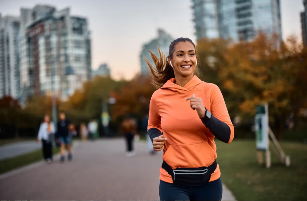 Smiling woman in an orange hoodie jogging in a city park with earbuds and a fitness belt, representing an active and healthy lifestyle.