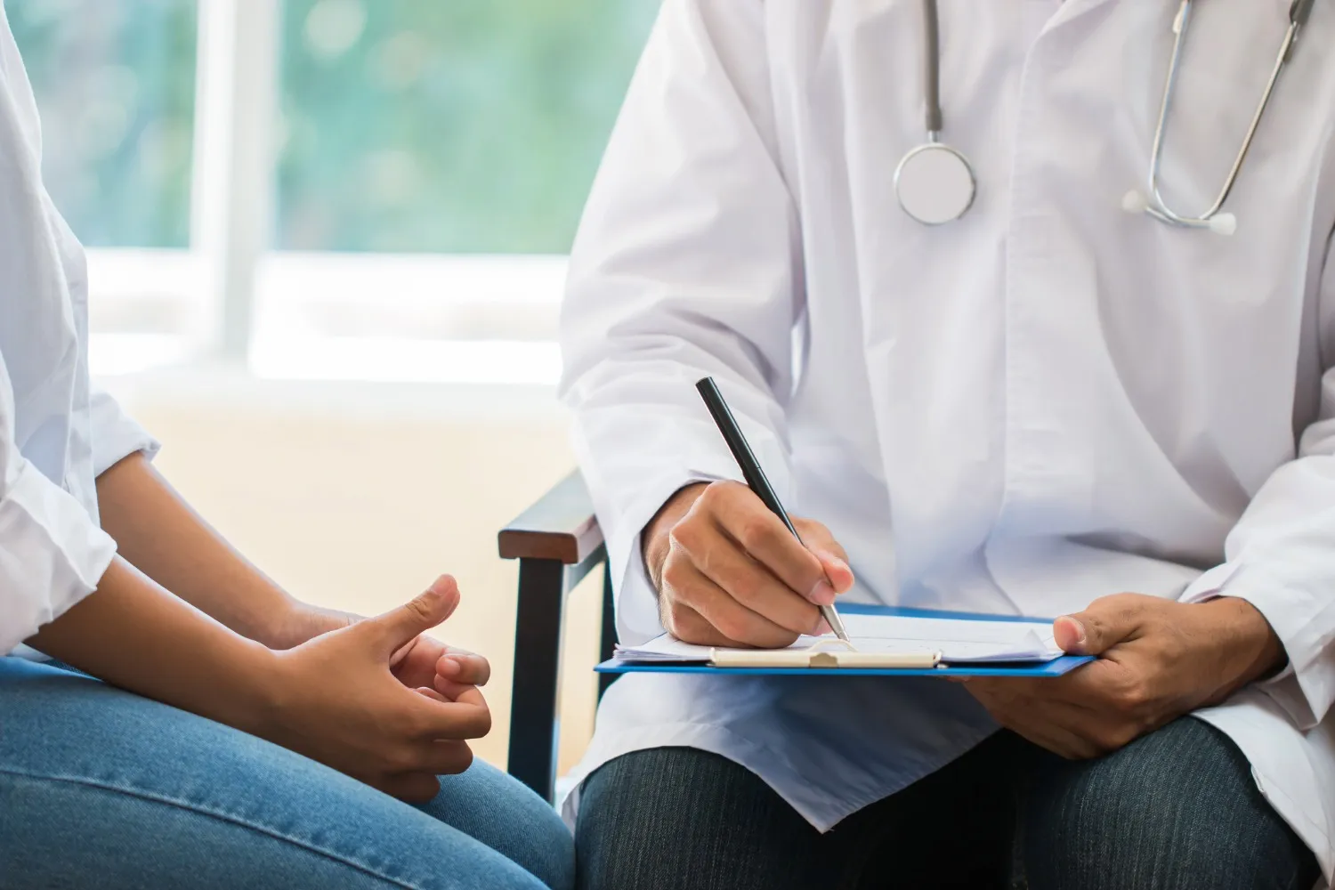 A doctor in a white coat with a stethoscope takes notes on a clipboard while speaking with a patient, who is sitting across with hands clasped; both are seated indoors during a medical consultation.