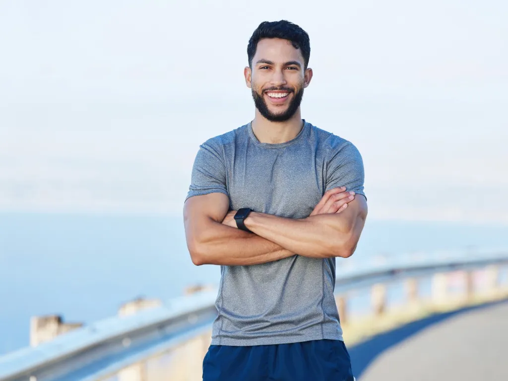 Smiling young athletic man in gray t-shirt standing outdoors with arms crossed near a roadside guardrail.