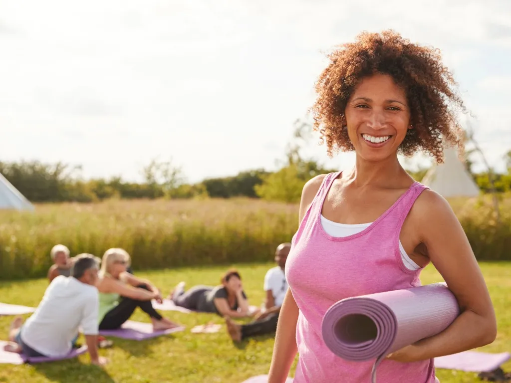 Smiling woman holding a rolled yoga mat outdoors with a group sitting on yoga mats in the background.