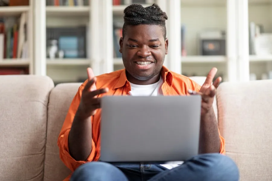 Smiling man wearing an orange shirt sitting on a beige sofa using a laptop in a living room.