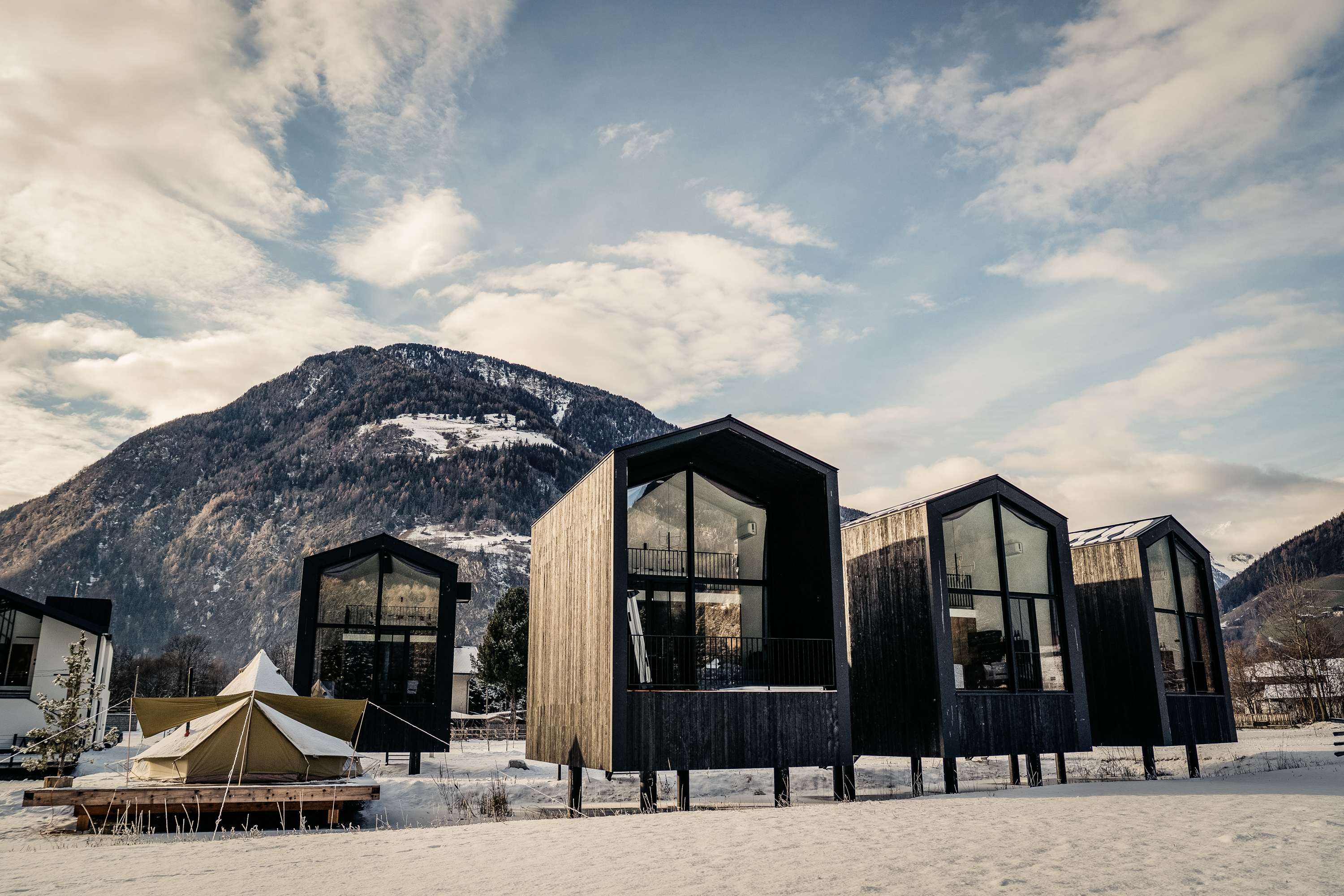 Modern black wooden cabins elevated on stilts in a snowy landscape with a green tent on a wooden platform and mountains in the background.
