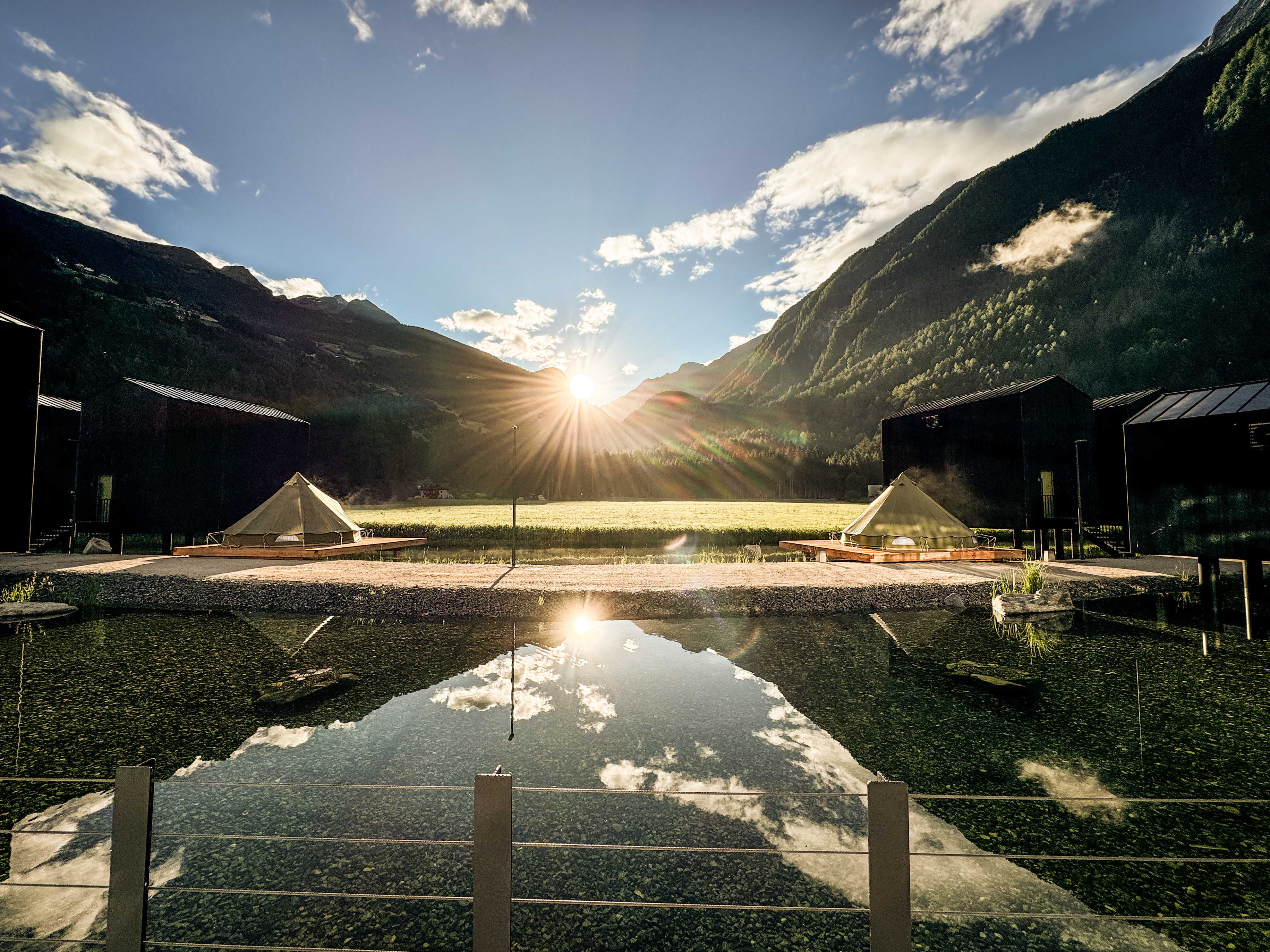 Sun setting behind mountains with two tents near a reflective pond and black cabins on either side.