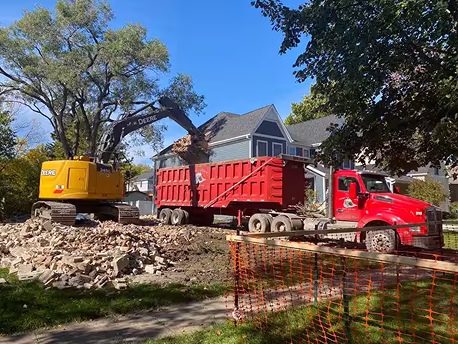 Excavator performing house demolition and loading debris into a roll off dumpster.