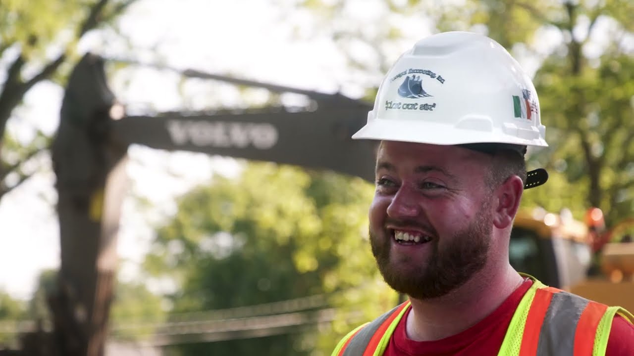 Smiling construction worker wearing a white hard hat and neon safety vest with blurry construction equipment and trees in the background.