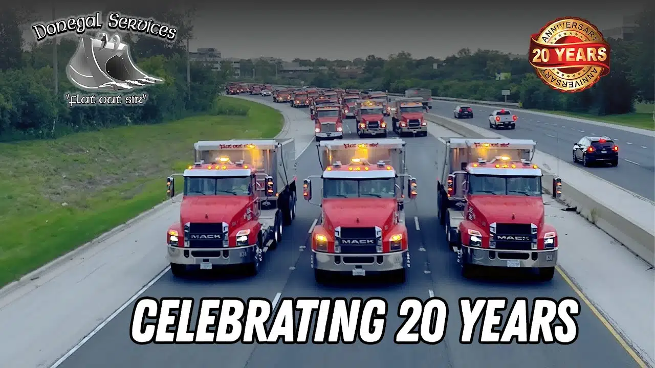 Three red Mack dump trucks driving side by side on a highway with many more trucks behind, celebrating 20 years with Donegal Services.