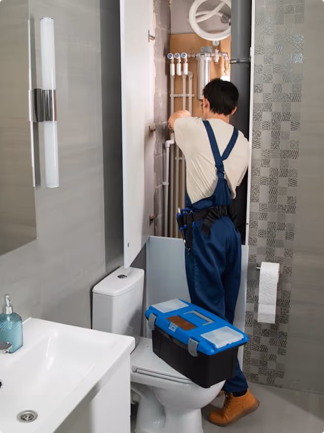 Plumber in blue overalls fixing pipes inside a bathroom cabinet above a toilet.