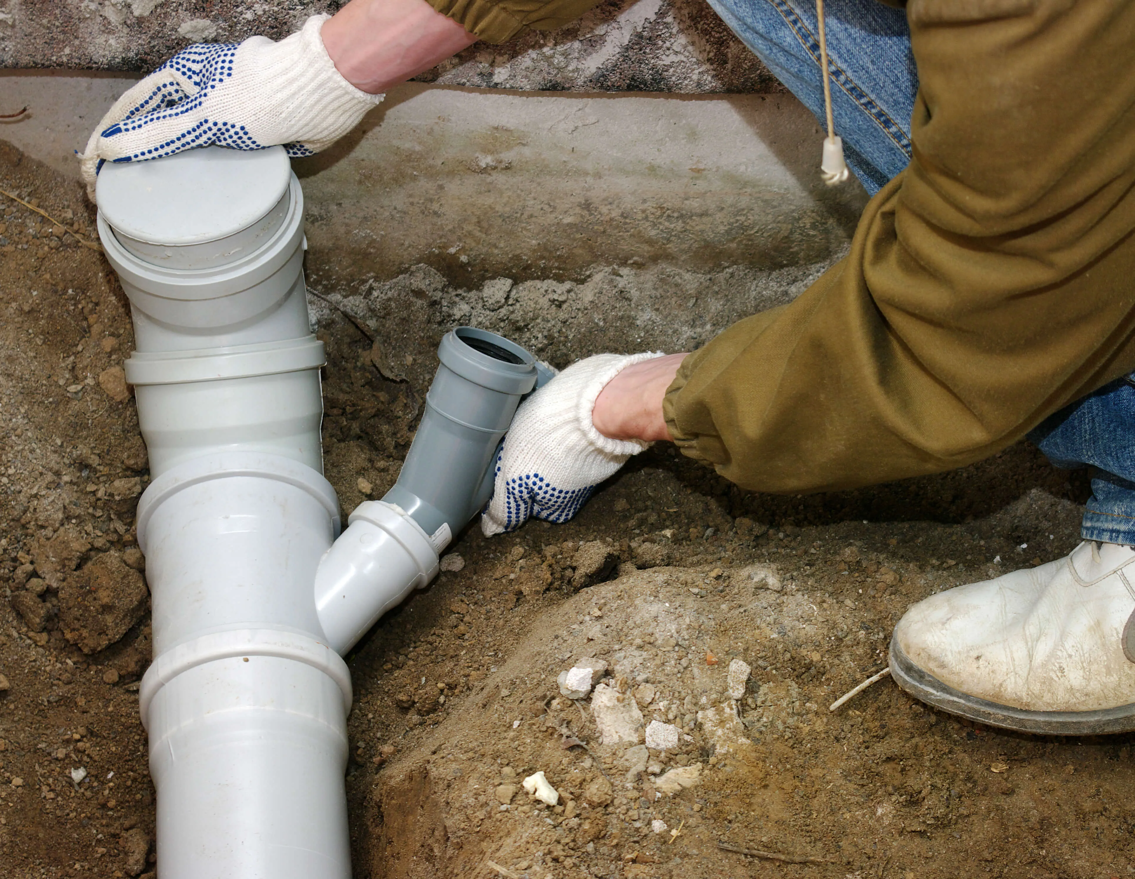 Person wearing gloves and work boots assembling white PVC sewage pipes underground.