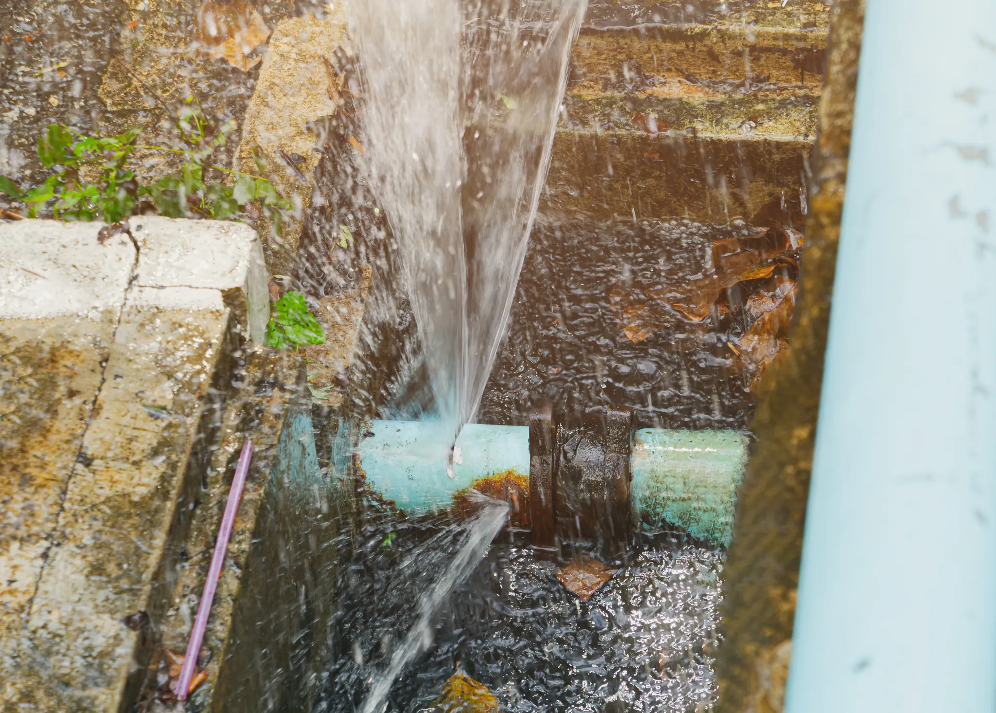Water bursting out forcefully from a broken turquoise pipe joint outdoors.