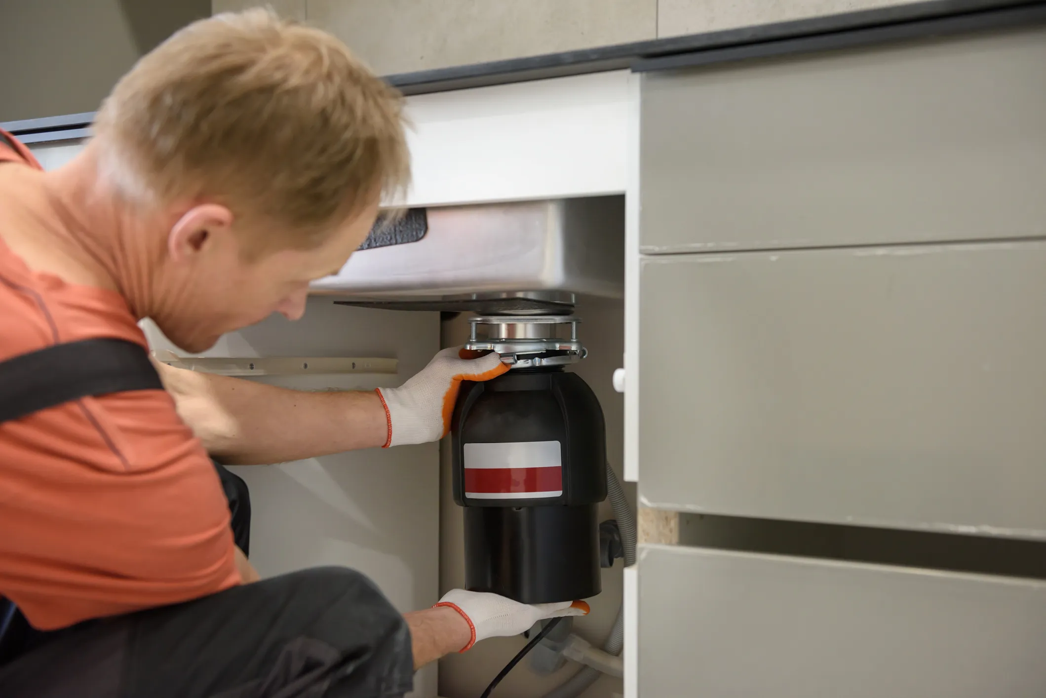 Technician installing a black garbage disposal unit under a kitchen sink.