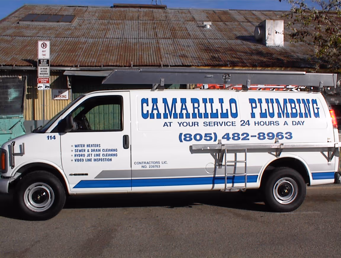 White plumbing service van with blue text reading 'Camarillo Plumbing, at your service 24 hours a day' and a phone number (805) 482-8963 parked in front of a rustic building.