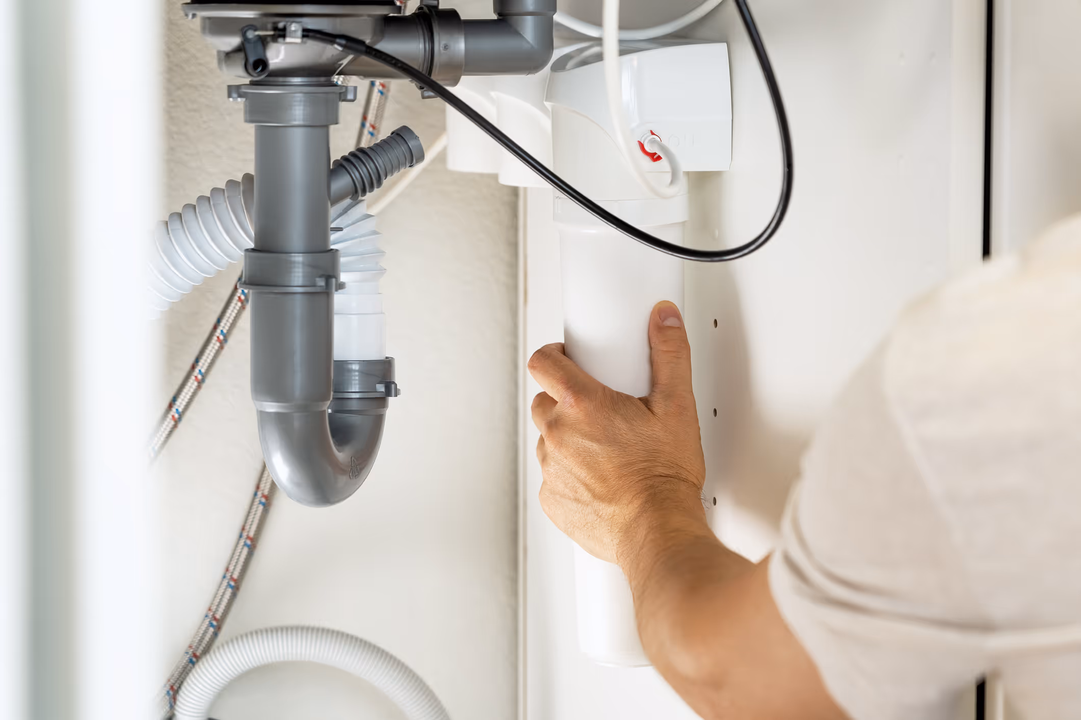 Person installing or adjusting a white water filtration or softener system under a sink with visible gray plumbing pipes.