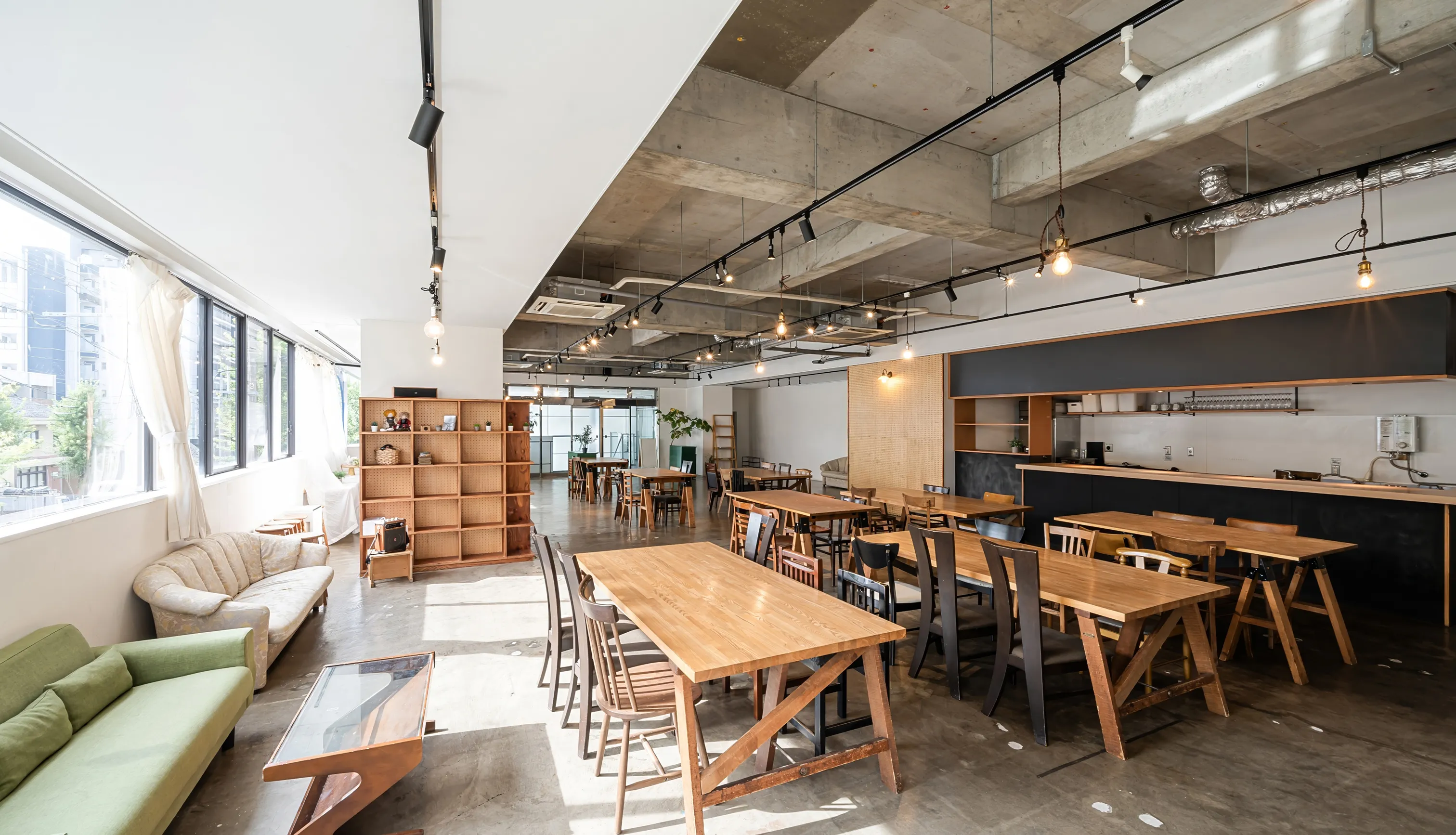 Modern open workspace with wooden tables, mixed chairs, large windows, and industrial ceiling lighting.