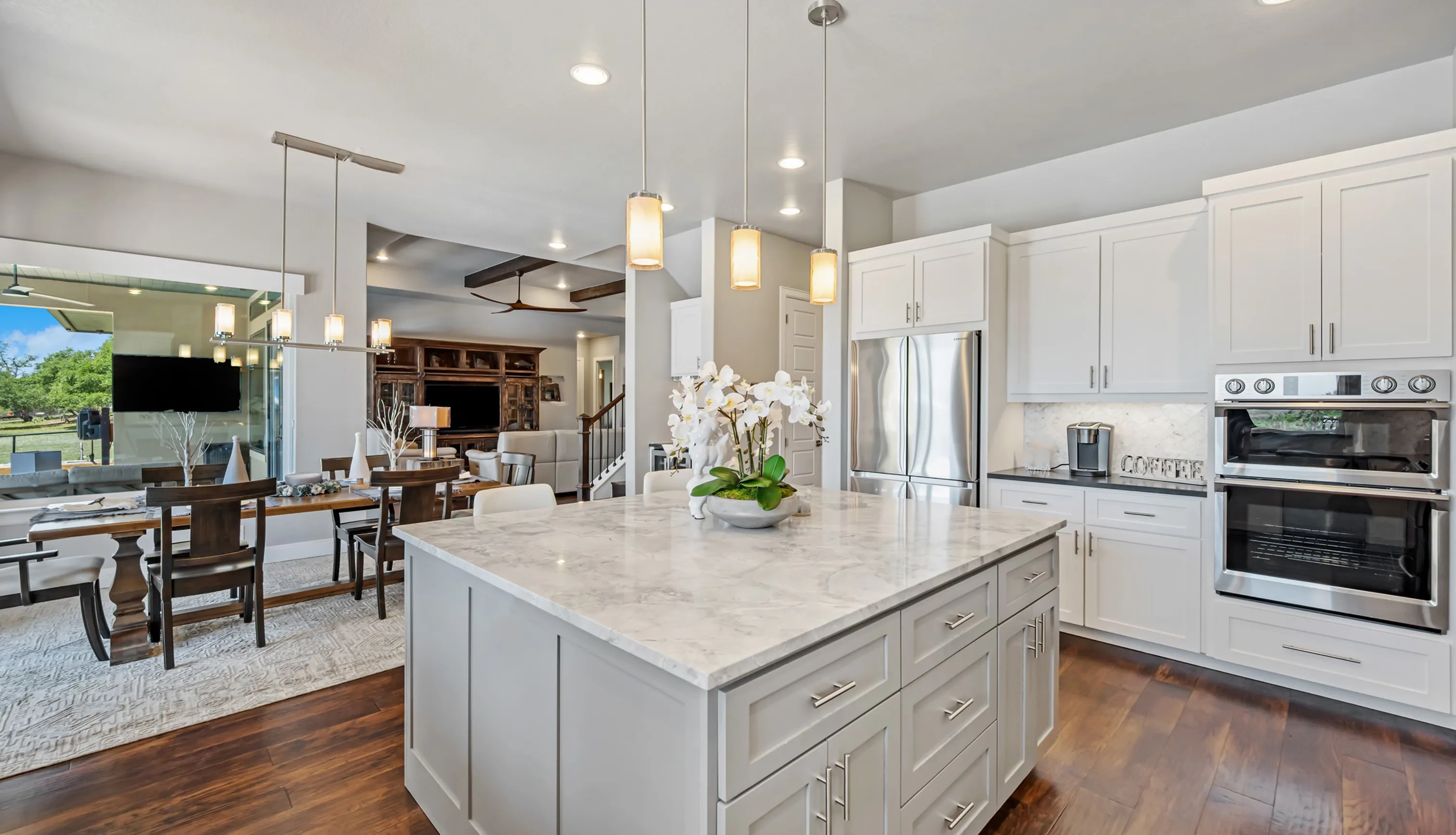 Modern kitchen with white cabinets, marble island with orchid centerpiece, stainless steel appliances, and open view to dining and living areas.