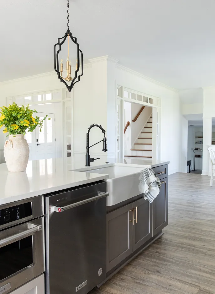 Modern kitchen island with a white farmhouse sink, black faucet, stainless steel dishwasher, and a vase of yellow flowers on a white countertop.