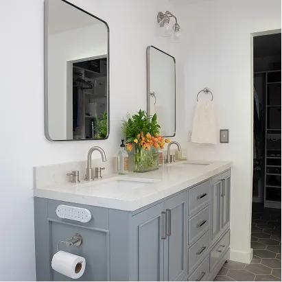 Modern bathroom vanity with double sinks, gray cabinets, two mirrors, a vase of flowers, and a towel with toilet paper holder on the side.