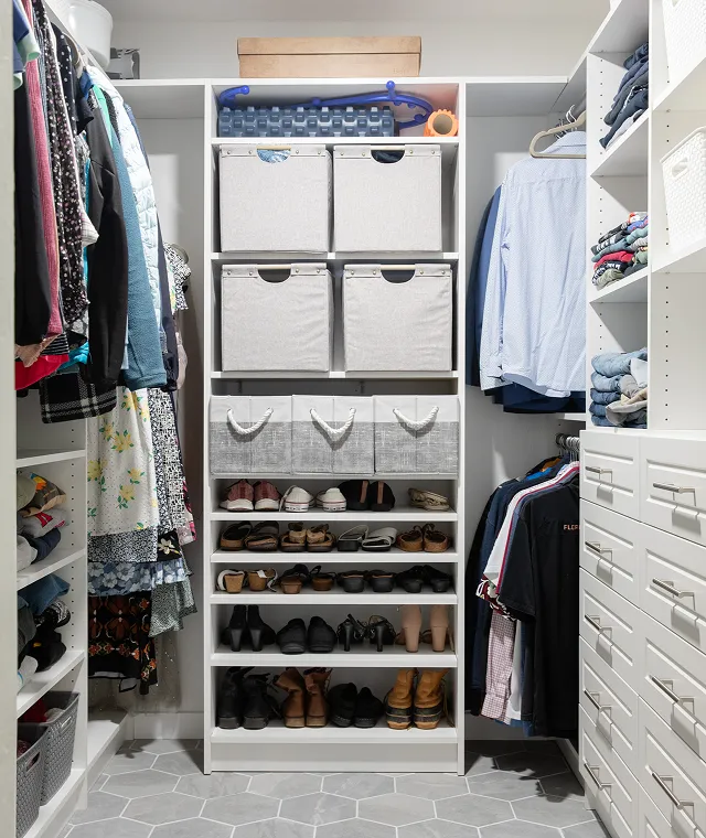 Organized walk-in closet with shelves holding fabric storage bins and shoes, surrounded by hanging clothes on both sides.