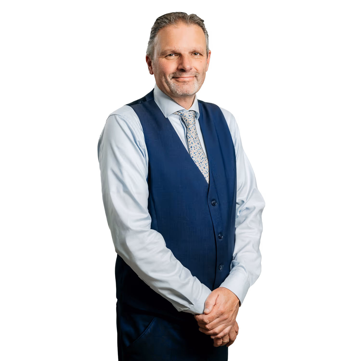 Zelfverzekerde man met lichte overhemd, blauwe vest en bloemenstropdas, poseert glimlachend met gevouwen handen.