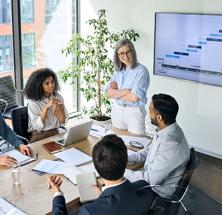A diverse group of professionals gathered in a meeting room, focused on a laptop in the center of the table.