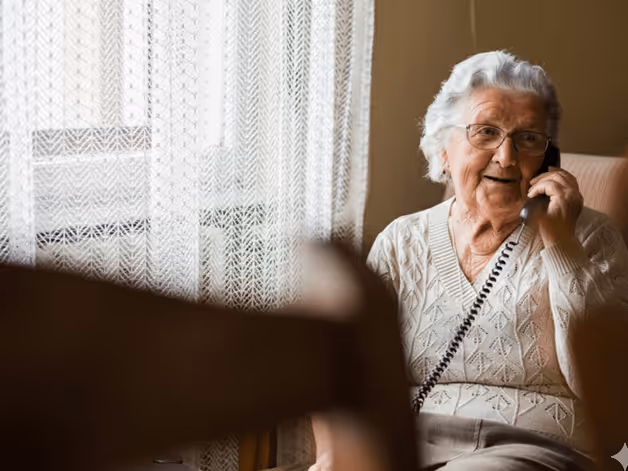 An elderly woman sits in a chair, engaged in a conversation on the phone.