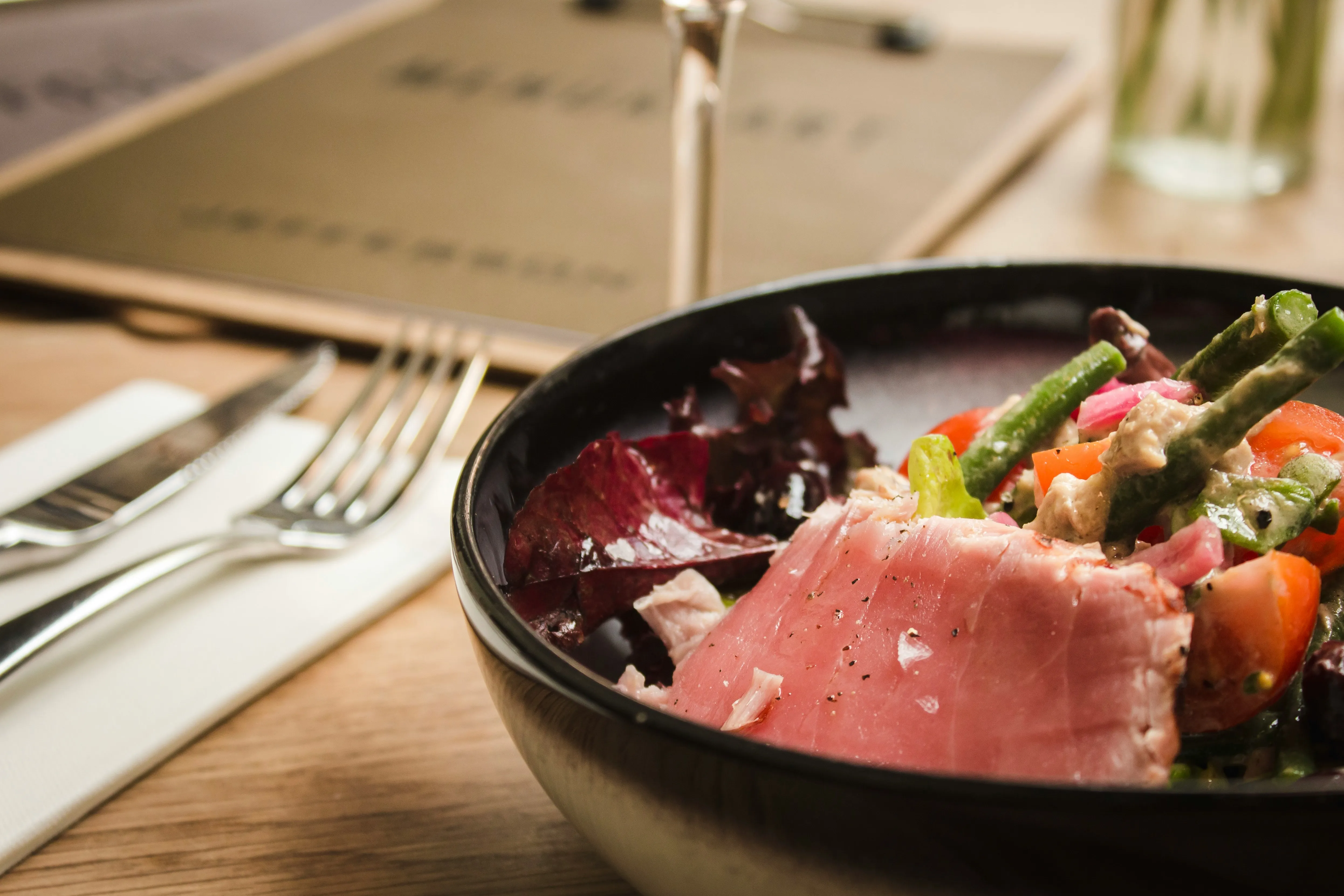 Close-up of a black bowl with fresh mixed salad containing lettuce, green beans, tomatoes, and a slice of seared tuna, on a wooden table with a knife and fork on a napkin beside it.
