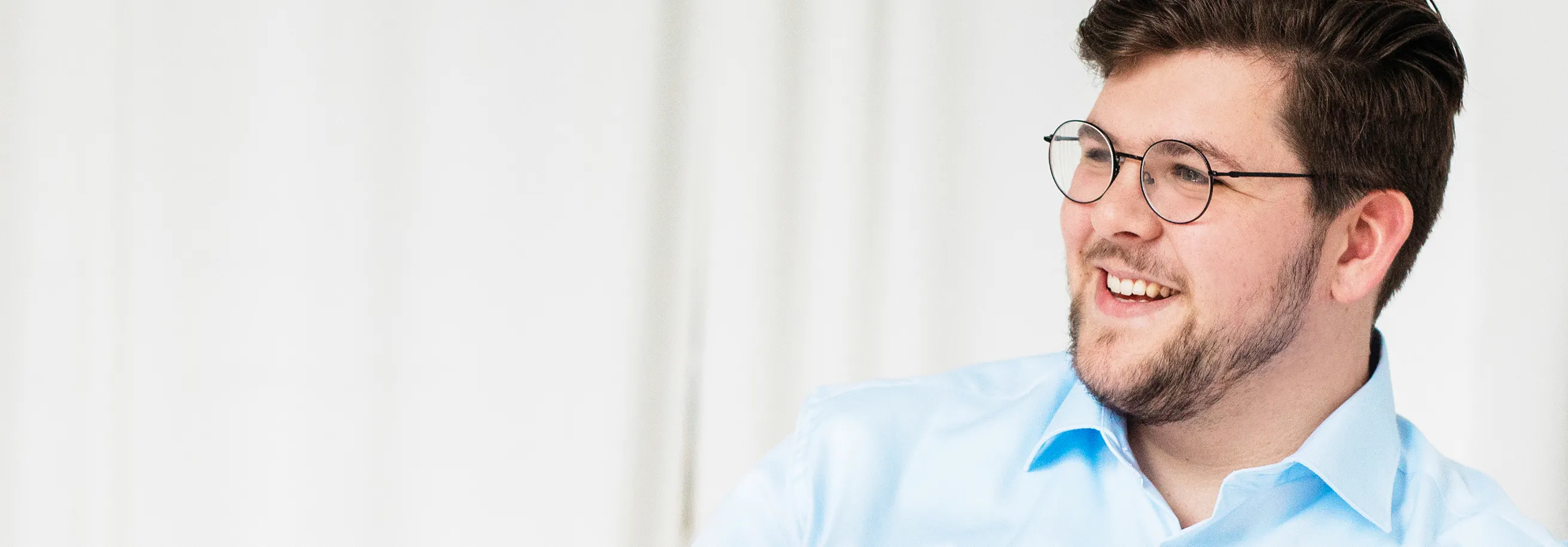 Young man with dark hair and beard wearing round glasses and a light blue shirt smiling and looking to the side.