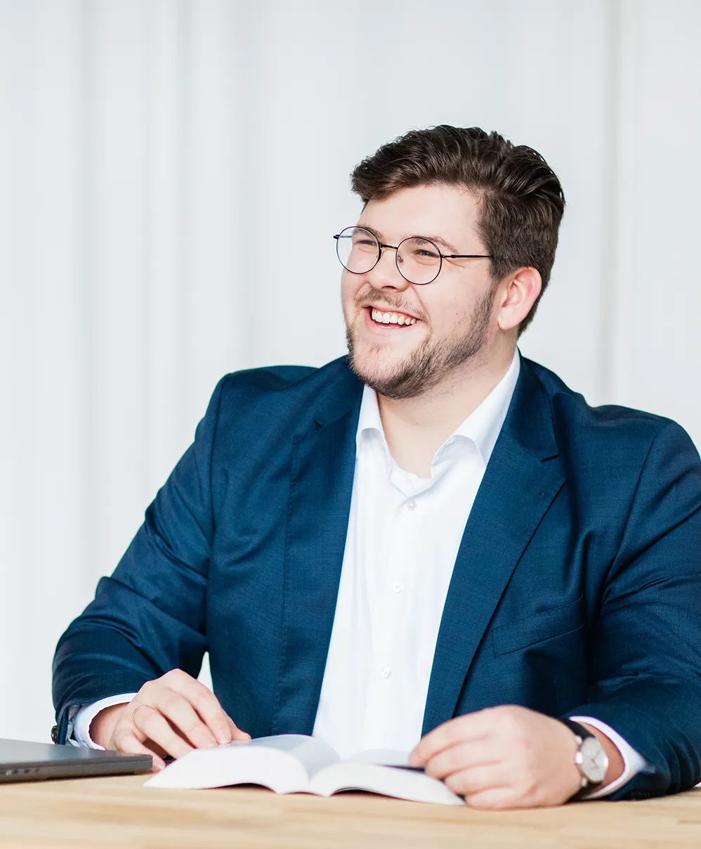 Smiling man with glasses and beard wearing a blue suit jacket and white shirt, sitting at a table with an open book and laptop.