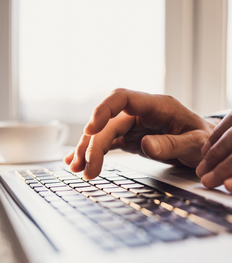 Close-up of a person's hand typing on a laptop keyboard with a white cup in the background.