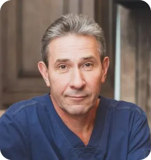 Middle-aged man with short gray hair wearing blue medical scrubs in a professional indoor setting.