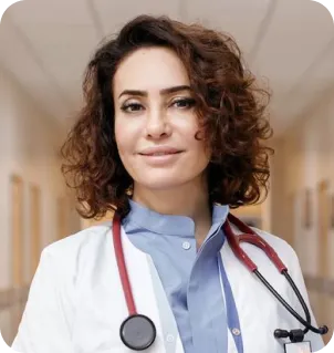 Smiling female doctor with curly hair wearing a white coat and red stethoscope in a hospital hallway.