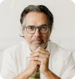 Middle-aged man with gray hair and beard wearing glasses and a white shirt, resting his chin on clasped hands.