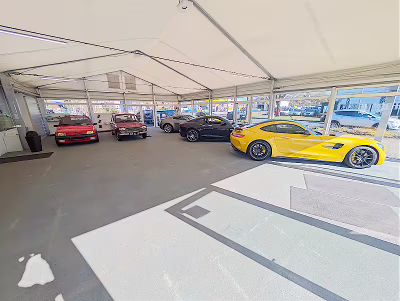 Vier geparkte Autos in einem hellen, überdachten Raum mit großen Fenstern, darunter ein gelbes Sportauto und zwei klassische Wagen.