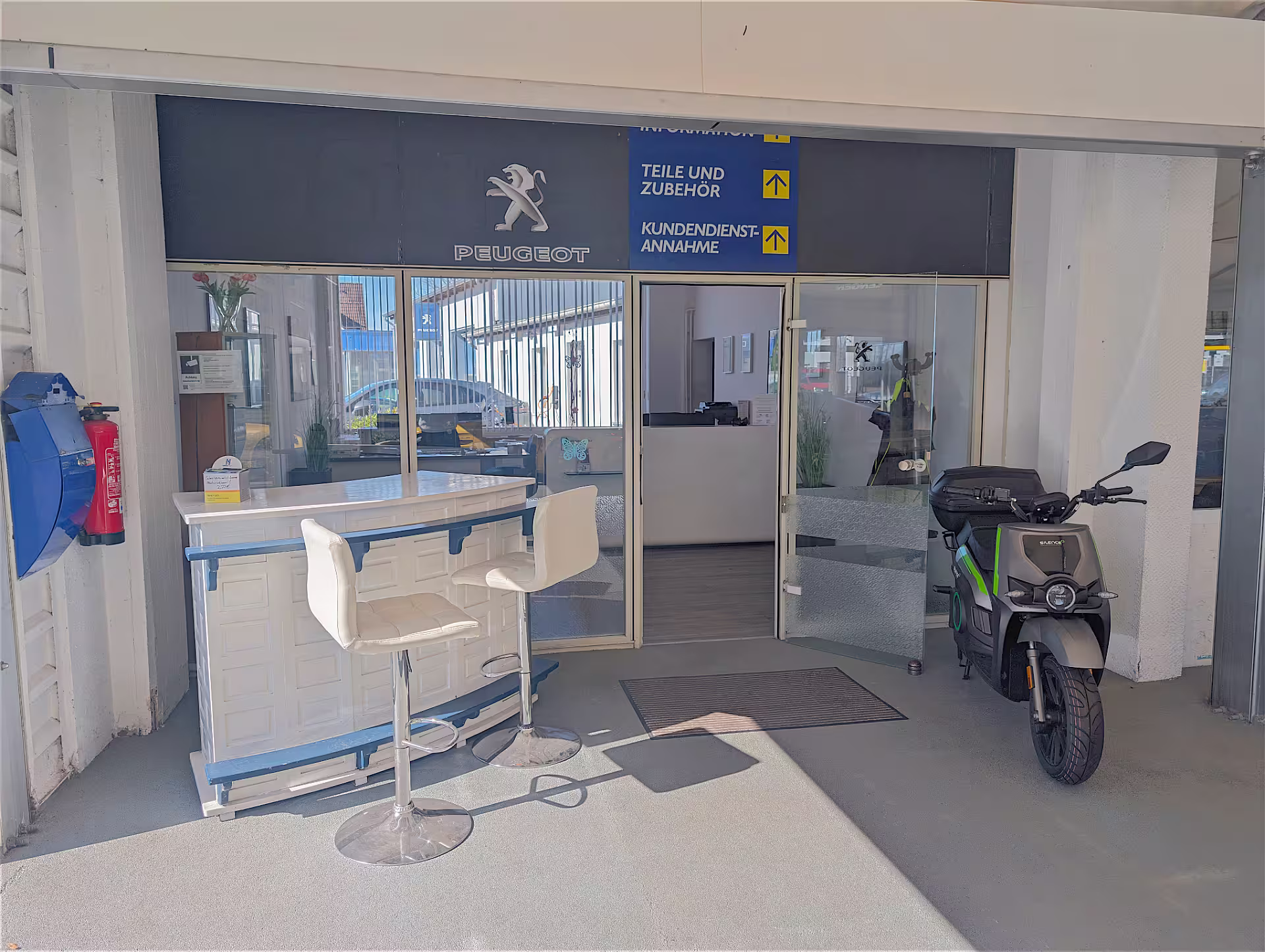 Fahrzeugempfangsbereich bei Peugeot mit zwei weißen Barhockern vor einem Empfangstresen und einem geparkten schwarzen E-Roller.