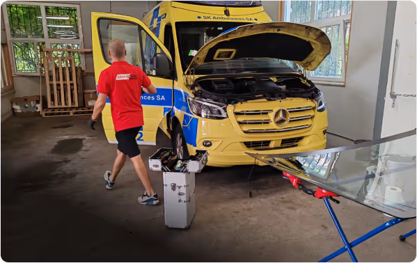 Un homme en t-shirt rouge marche près d'une ambulance jaune avec le capot ouvert dans un garage.