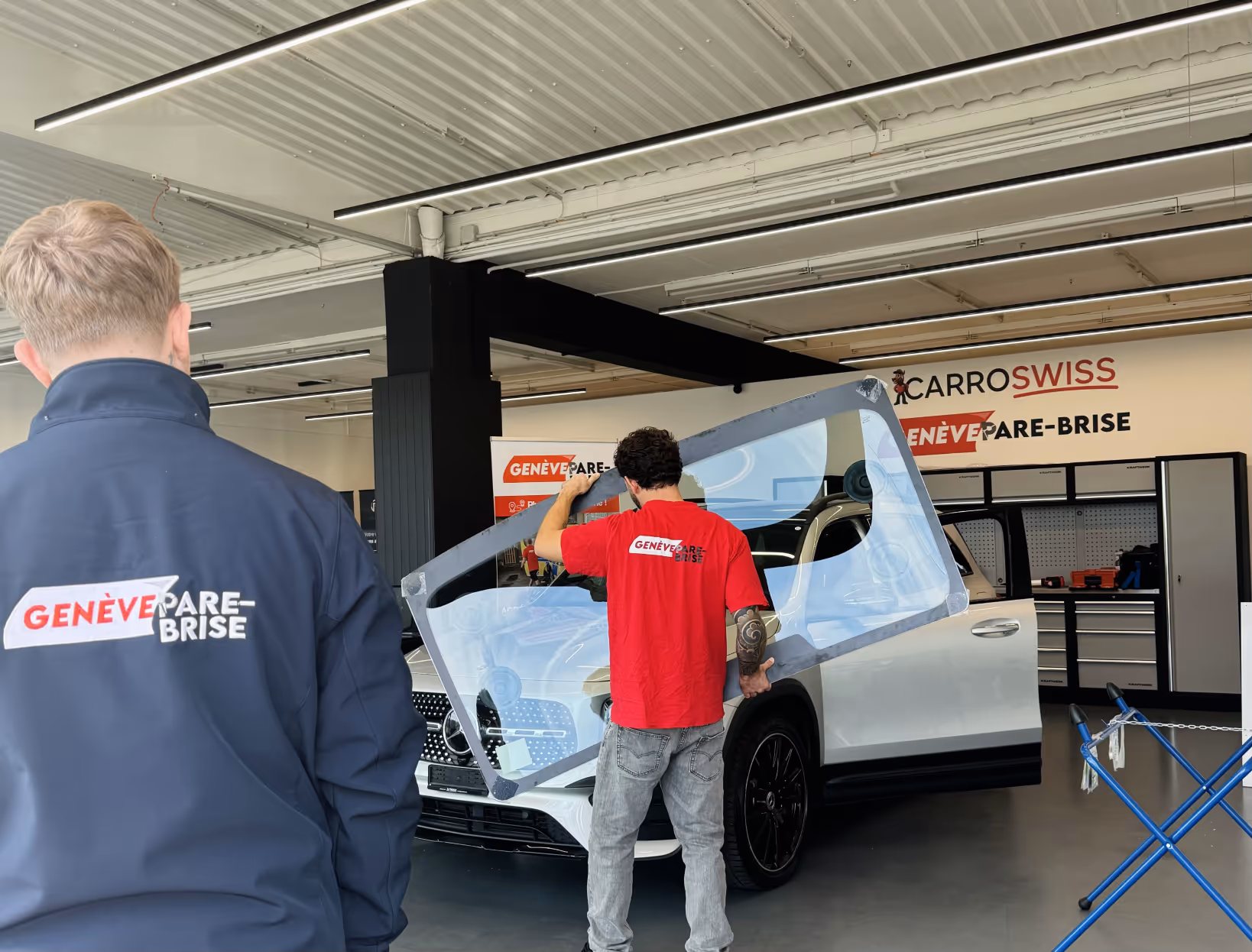 Deux techniciens Genève Pare-Brise dans un atelier, l'un tenant un pare-brise prêt à être installé sur une voiture blanche.