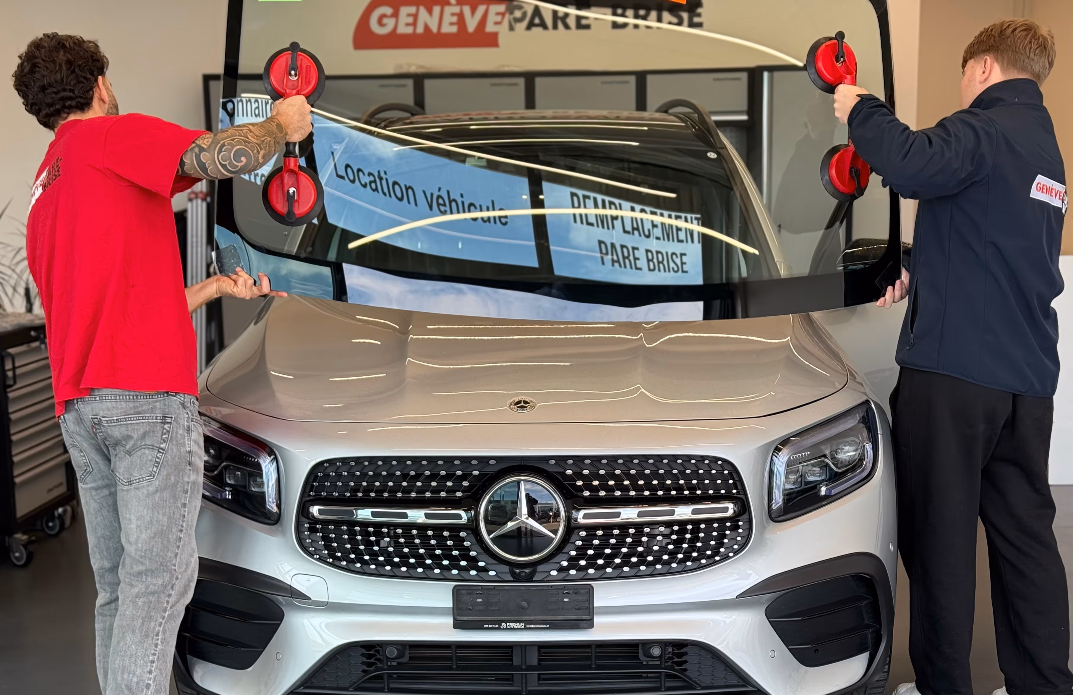 Deux hommes installant un pare-brise neuf sur une Mercedes blanche dans un atelier.
