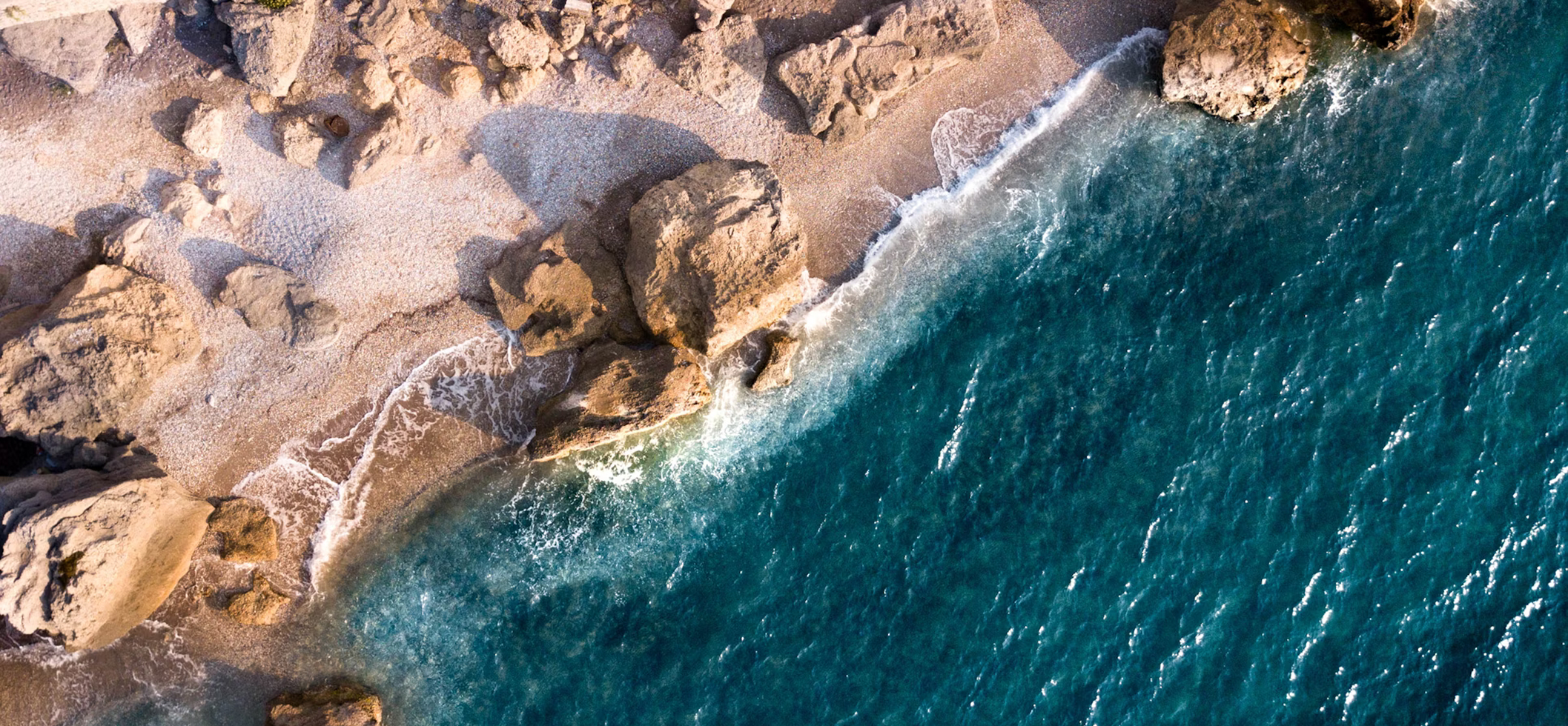 Luftaufnahme von Wellen, die an felsige Küste mit sandigem Strand schlagen.
