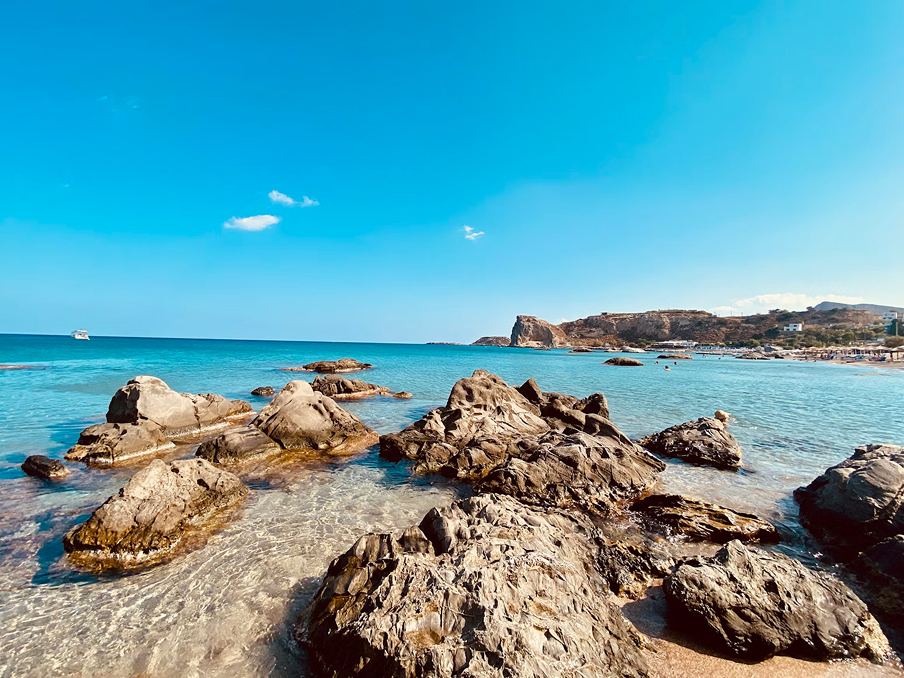 Klarer blauer Ozean mit großen Felsen im flachen Wasser und felsige Küste im Hintergrund unter blauem Himmel.