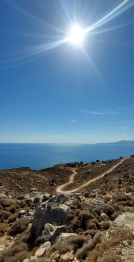 Heller sonniger Himmel über einer kurvigen Straße durch eine trockene, felsige Küstenlandschaft mit Blick auf das blaue Meer.