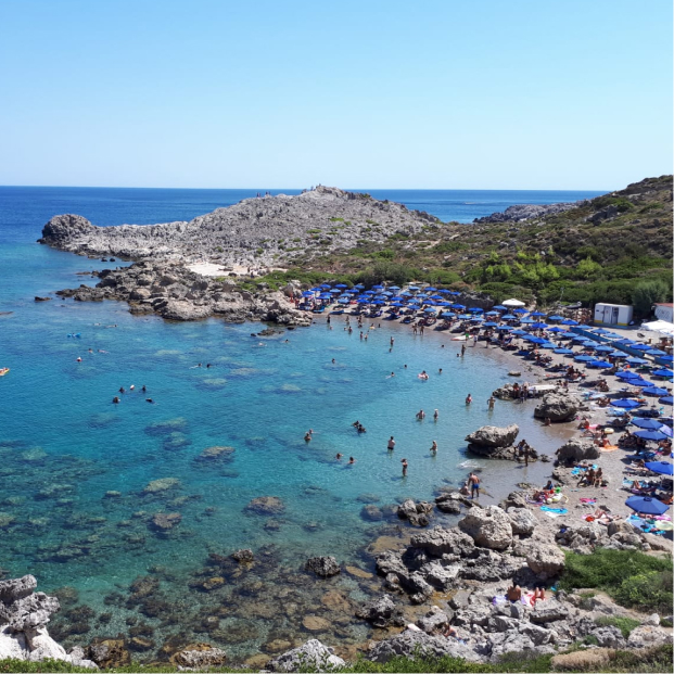 Felsenstrand mit klarem blauem Wasser, vielen Badegästen und blauen Sonnenschirmen unter blauem Himmel.