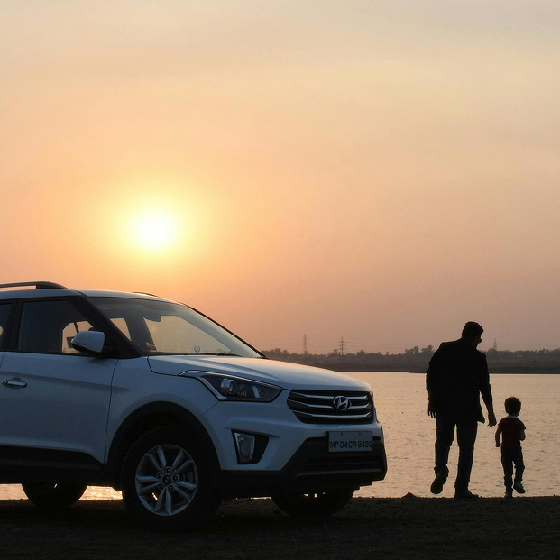 Ein weißes SUV steht am Ufer eines Gewässers bei Sonnenuntergang, während ein Erwachsener und ein Kind nebeneinander am Wasser entlanggehen.
