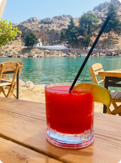 Ein roter Cocktail mit Limettenscheibe und schwarzem Strohhalm auf einem Holztisch am Meer auf Rhodos mit Bergen und Bäumen im Hintergrund.