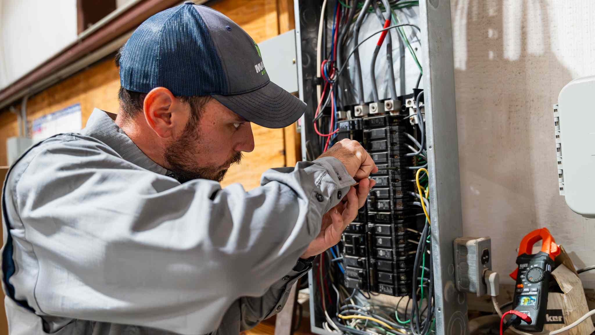 Electrician in a cap and gray shirt working on an electrical breaker panel with wiring and tools nearby.