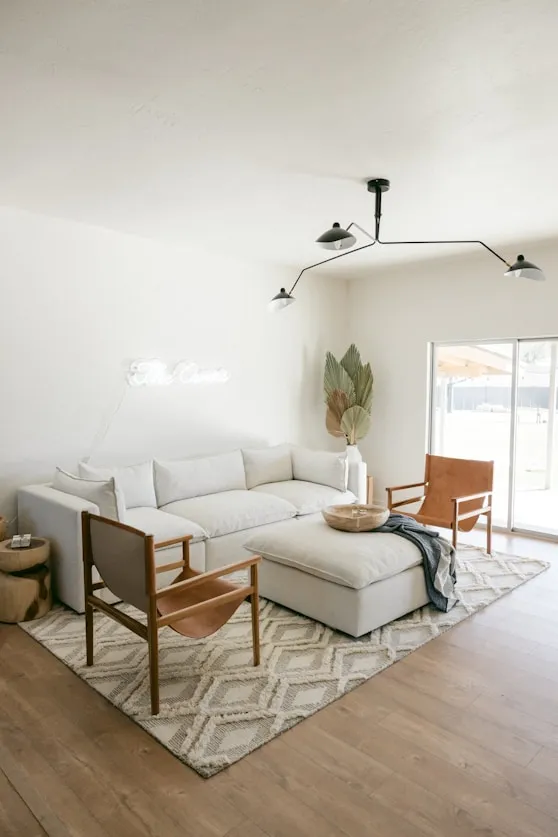Minimalist living room with a large white sofa, two tan leather chairs, a patterned rug, and a modern black ceiling light fixture.