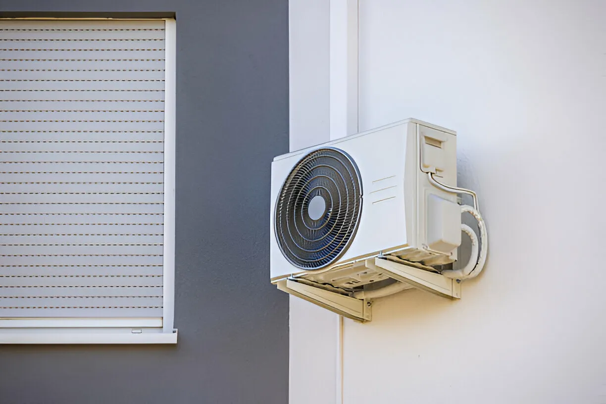 Outdoor unit of a white air conditioning system mounted on a wall next to a closed window with a white shutter.