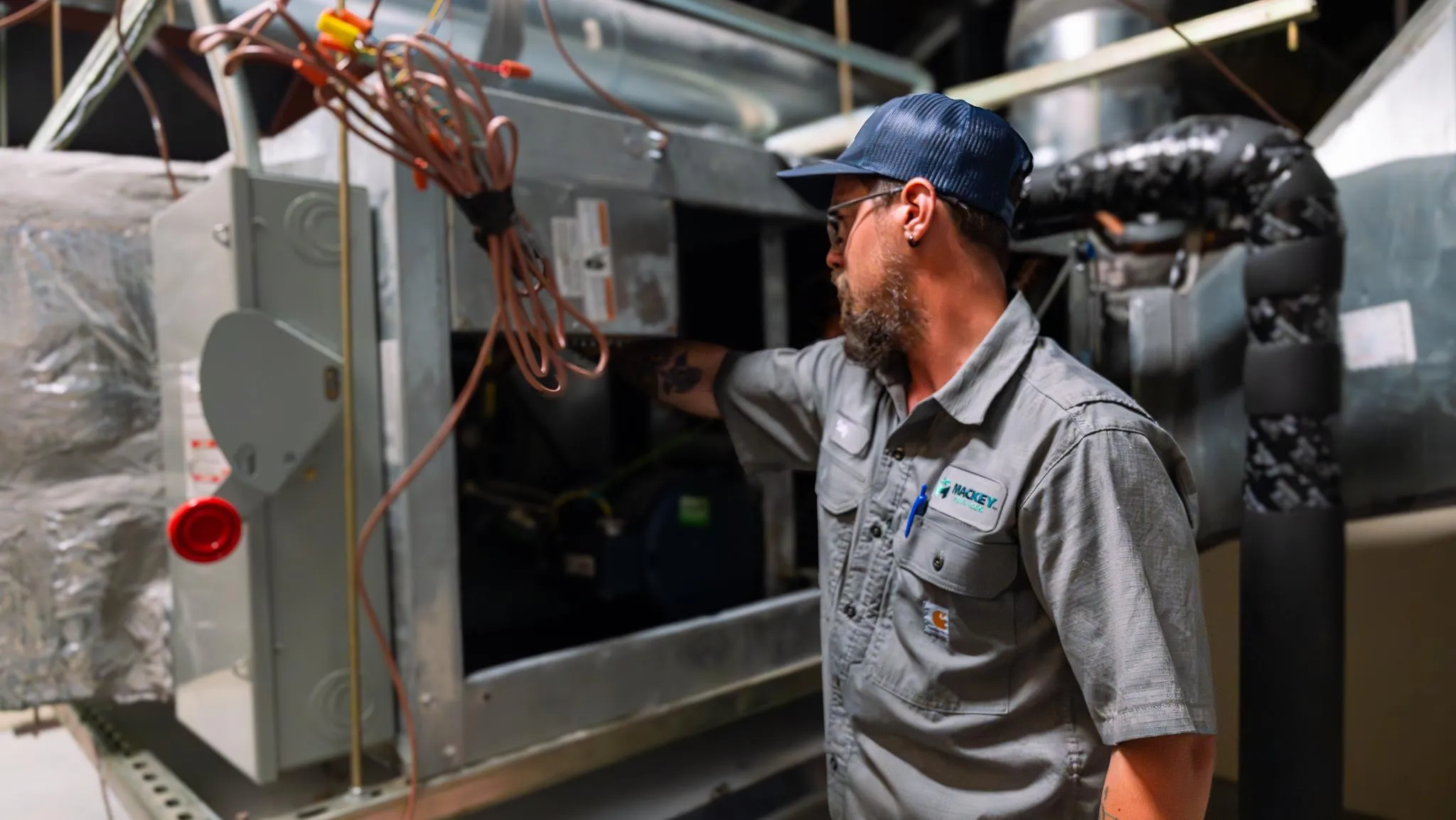 Technician wearing a cap and glasses inspecting an open industrial HVAC unit in a utility room.